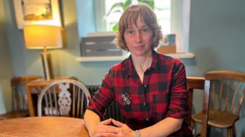 Gemma Williams is sat at a table looking at the camera. She is wearing a red checked shirt with her sleeves rolled up and has short brown hair with a fringe. Chairs and a radiator can be seen in the background. 