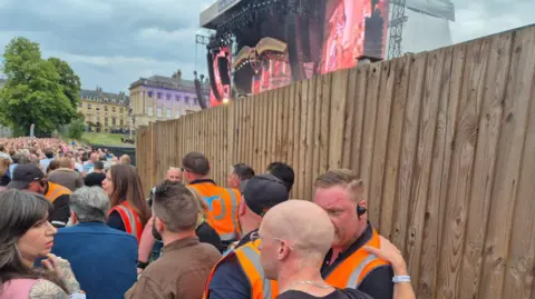 Claudine Beard A picture of a crowd of people at a concert with a number of security guards wearing orange jackets. There is a large wooden fence blocking the stage with only a large screen and the roof of the stage visible. 