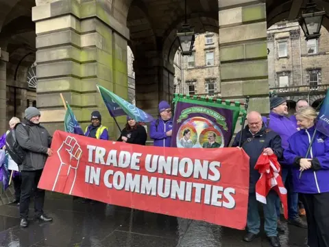 A group of people, mainly men, holding a Trade Unions in Communities banner in front of the columns of an old building