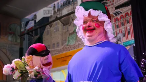 A man is dressed like a pantomime dame with a white lace frilly mop cap, a green and red baseball cap on top, pearl necklace and a blue top. His make-up is over the top with exaggerated red blusher and red lips. He is holding a puppet and a bunch of artificial flowers