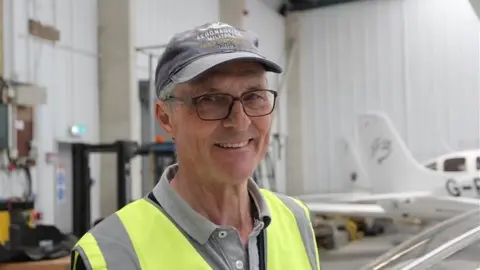 A man in a cap and hi-vis jacket standing inside a warehouse building where aeroplanes are kept.