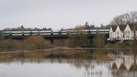 PA Media A green, white and yellow train crosses a bridge above a flooded river with three white houses on the right side riverbank over a