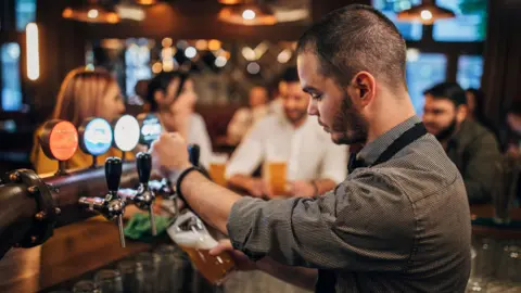 A bartender, with short black hair and a black stubble, wearing a grey shirt, standing behind a bar. He is pulling a pint of beer. There are four people in the background, which is blurred.