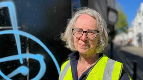 Emma Humphreys standing in Trafalgar Street, Brighton, next to a wall with blue spray paint on it. She has grey hair and is wearing a high vis vest