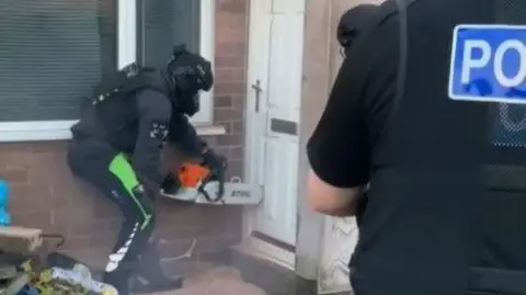 A police officer wearing protective clothing uses a chainsaw to cut through the front door of a house, while another officer stands watching.