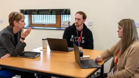 Northamptonshire Police, Fire and Crime Commissioner Three people in a room, all talking to each other, sat round a table. A woman, on the left, has short hair with glasses on her head, has one arm up and is looking across a table at another woman. She is wearing a grey jacket, with a folder in front of her. A man, next to her is looking at her, with a black hoodie on, with a lanyard round his neck. He has a laptop in front of him. He has short hair and a beard. The woman to the right, has long blonde hair, has a laptop in front of her and is wearing a beige top, There is a window in the room with a blind at it. 