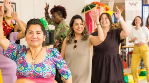 Three women raising their hands in a line wearing a dress.  There are women behind them in a circle raising their hand. 