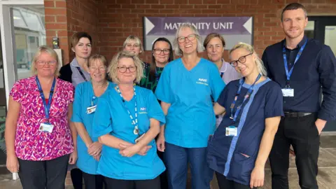 Somerset NHS Foundation Trust Sue Lee, pictured in the centre of a group of colleagues, all of whom are smiling at the camera. She has short white hair, cut in a bob, and dark round glasses. She wears her uniform of a bright blue short-sleeve tunic and dark blue trousers. The group are outside the maternity unit of the hospital. 