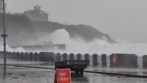 Waves crash onto the shore at Onchan Head, there is a road closed sign in the foreground and debris across the road. 
