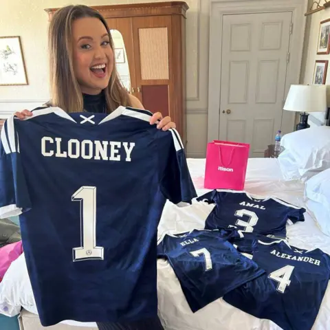 Claire Phale A woman holding up a navy Scotland football shirt with “Clooney” printed on the back, smiling in a hotel room with gift bags and folded shirts on the bed.