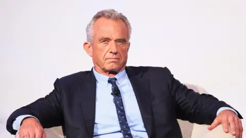 RFK Jr, wearing a suit, sits in an arm chair with his arms on the sides, with a neutral expression