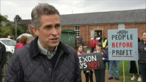 A man wearing a coat stands outside speaking to the camera. People are stood behind him holding placards and signs.