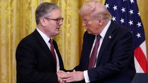 Sir Keir Starmer and Donald Trump stand facing each other, shaking hands. Both are dressed in dark suits, white shirts, and red ties. Behind them is a yellow curtain and a prominently displayed American flag.