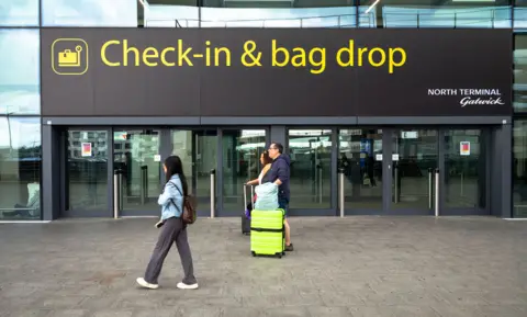 Universal Images Group via Getty Images A sign outside Gatwick airport reads 'check-in & bag drop; north terminal Gatwick', while people walk past wheeling baggage along the concrete floor, image taken in 2024.