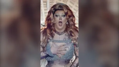 Kate Butch looks shocked with her hand covering her chest. She has long wavy ginger hair and wears a knitted bikini top.