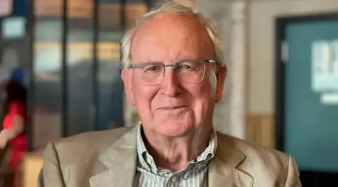 Tony Beckingsale is smilling in the photo. He is wearing glasses and has grey hair. He is also wearing a striped shirt and a blazer.