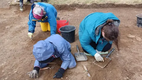 Three people are on their knees and crouched over as they investigate the soil below them. They are knelt in a shallow hole that has been dug in the field. They are all wearing blue waterproof jackets and gloves while archaeology equipment like spades and spatulas are strewn around them.