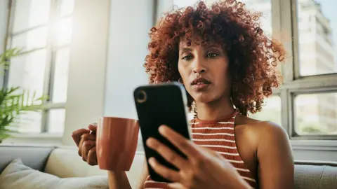 A woman looks at her phone with a mug in hand inside her home