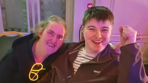 Caring Breaks Two women, cheering and smiling at camera. They both have brown hair. The woman on the left is wearing a black hoody and has a neon glowstick attached. The woman on the right is wearing a brown hoody and striped white top. They are sat at chairs with neon lights visible.
