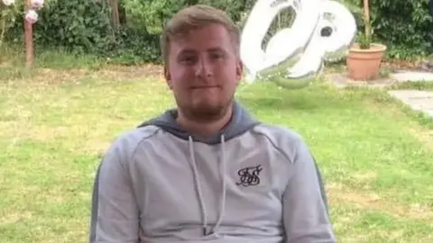 David Johnston Bristow smiles as he sits in a garden wearing a grey tracksuit top.