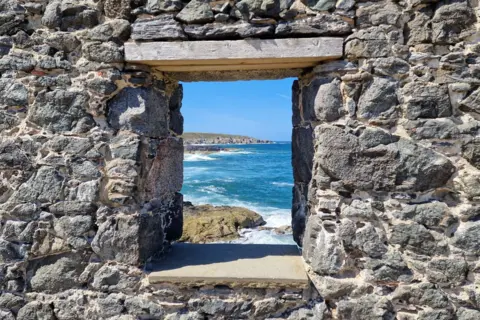Matthew Jackson A rectangular hole cut through a rocky wall reveals a coastal scene on the other side with the bright blue sea creating foam as it hits against rocks.