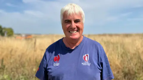 Tim Carson is standing in a field wearing a blue T-shirt. He has short, white hair and is smiling.