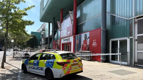 An external view of the store, with red and white signage and the Arsenal logo. A police car is parked outside and police tape has been installed.