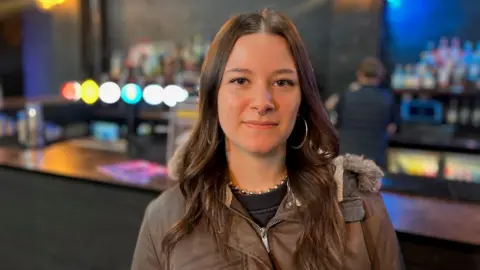 A woman wearing a brown jacket is standing in a bar. She has brown hair and a nose ring as well as large hoop earrings.