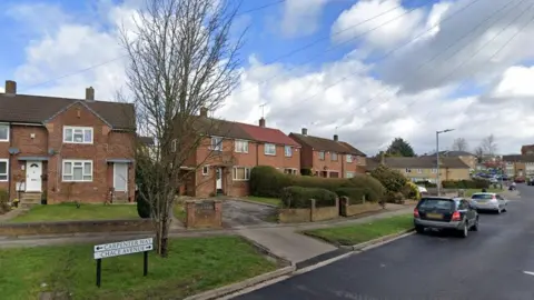 Google A residential street, with brick two-storey terraced housing. The houses have white window and door frames, and small lawns in front of them. There are low brick walls separating the gardens from the pavement. There are grass verges on the pavement and cars parked on the road. There is a street sign showing Carpenter Way to the left and Chace Avenue to the right. 
