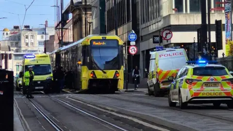 A tram stopped in the middle of the road, the front panel is missing. A police van and car are parked on the right side of the road. An ambulance is stopped next to the tram on the left. An officer wearing hi vis is stood in front of the ambulance.