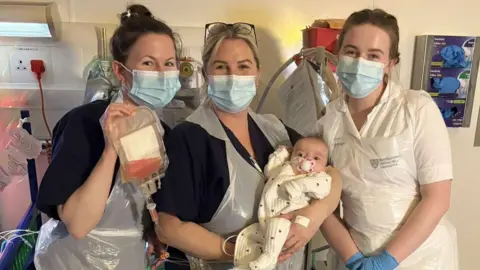 Family photo Dolcie-Mae has wispy brown hair, a red bow on her head and a white baby grow with love hearts on it. She is being held by three medical staff members at the hospital who all have protective masks on and white aprons. One of them is holding a blood bag.