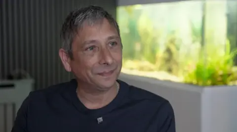 Nicole's father Jason sits in front of a fish tank. He has short grey hair and is wearing a black t-shirt.