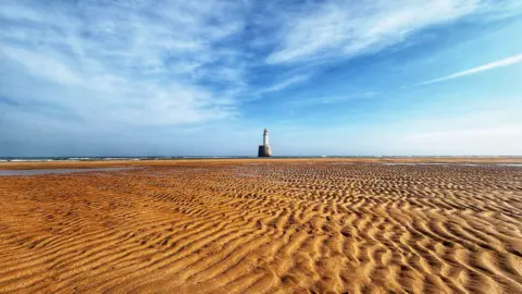 ALANA WILLOX A beach with tidal lines. The sand is golden. The sky above is mainly blue with white cloud marks. In the centre of the image in the distance is a white lighthouse with a red top.