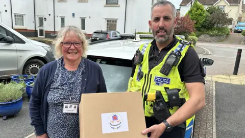 Mrs Wade and Sgt James Gallienne are holding a compassionate box. They are standing outside. Sgt James Gallienne is wearing a hi-vis police jacket. 