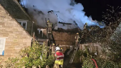 NFRS Thatched cottage - roof gutted by fire, smoke can be seen and four fire officers are on scene, three up ladders. All wear protective gear and helmets. 