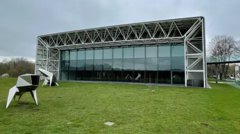 A big grey and white building fills up most of the picture. The front of the Sainsbury Centre is seen, with a massive glass window filling up the whole of the front of the building. There are two sculptures in the foreground.