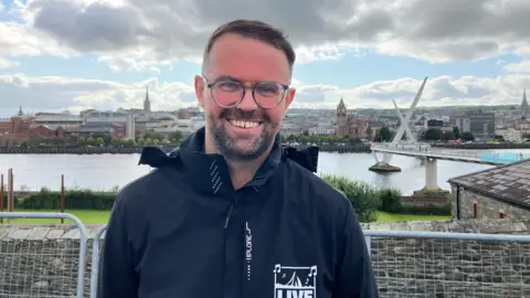 Niall O'Kane. He has short brown hair and beard, wearing glasses and a black raincoat with Live branding. The Derry cityscape is behind him.