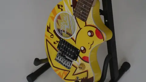 Shaun Whitmore/BBC The Fernandes Zo-3 Pokemon-themed limited edition electric guitar. It is yellow with the character Pikachu painted on it. It is sitting on a black stand.