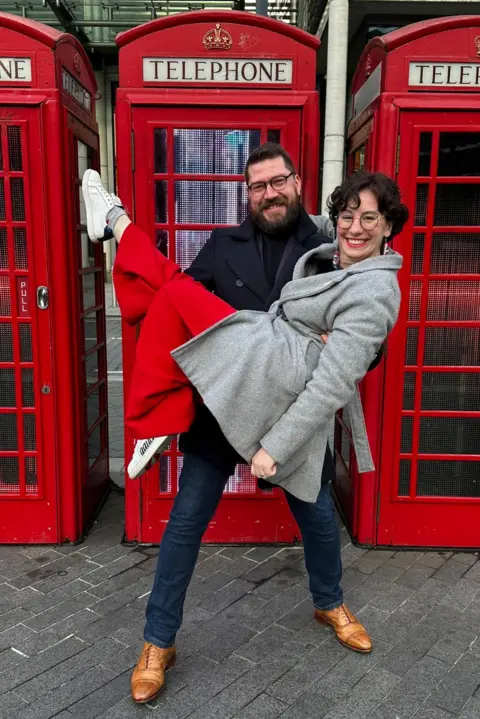 PA Media A man wearing a black jacket and blue jeans is holding a woman in front of a row of red telephone boxes. She is wearing a grey jacket and red trousers. They are both smiling.