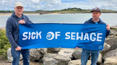 Two men, both wearing blue, stood by the sea holding a sign that says sick of sewage