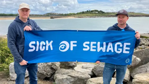 BBC Two men, both wearing blue, stood by the sea holding a sign that says sick of sewage