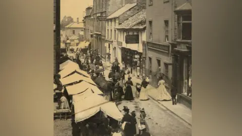 Keswick Museum A black-and-white photo from 1880 depicting several stalls with cloth awning on a cobbled pavement, while people who mostly seem to be women mill about them. Goods are being sold in the carts. There are old Victorian shop fronts in the background. The women are in Victorian-style dresses, with long billowing skirts, and some carry umbrellas.