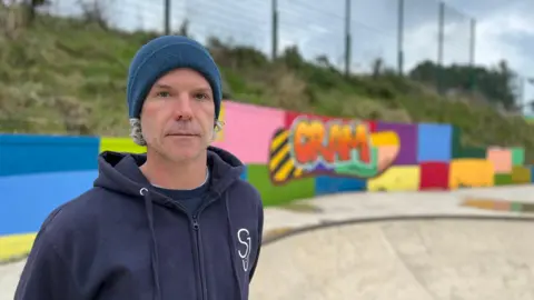 Phil looks at the camera wearing a blue hoodie with the letters SJ on his chest. He has a blue beanie hat with grey curly hair coming out of it. He is standing in a skate park with graffiti in Danny's memory behind him. 