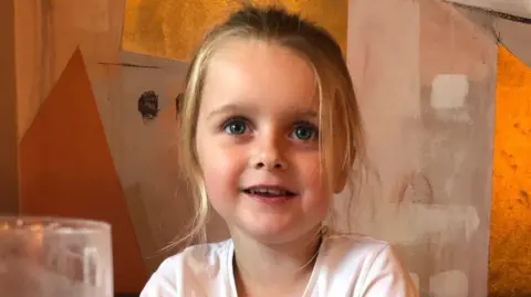 Bonnie Marsh has blonde hair in a ponytail and is sitting on a chair in front of a wall which is painted orange and brown. She is smiling at the camera.