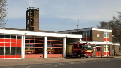 Humberside Fire and Rescue The exterior of a fire station. The large brick building has four large red and white doors containing fire engines. A further fire engine is pulling out onto the road.