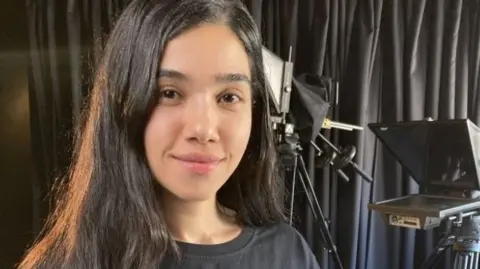 A girl with long dark-brown hair and wearing a black t-shirt standing in front of a production camera