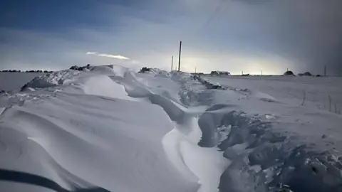 Ed Morrow Snow-covered road with deep drifts and power lines under a cloudy sky.