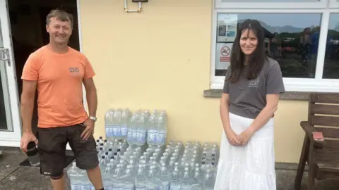 Victoria, wearing white skirt and grey top, and Trevor Vercoe in orange t-shirt, standing near plastic bottles of water.