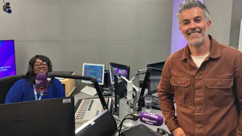 A woman, to the left of the image, sitting in a radio studio behind a microphone. She is wearing headphones and a blue jumper. A man is standing to her right. He is smiling and wearing a brown shirt.