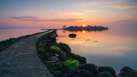 NOREENSIRELAND A stone pier leading down to still water. The sun is coming up in the horizon and the sky is pink, blue and peach. 